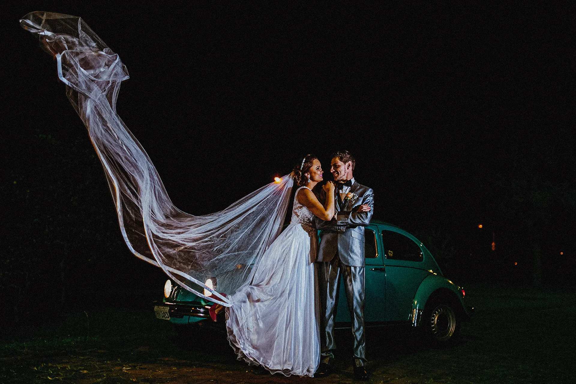 Casal ao lado de um carro clássico com o véu em movimento